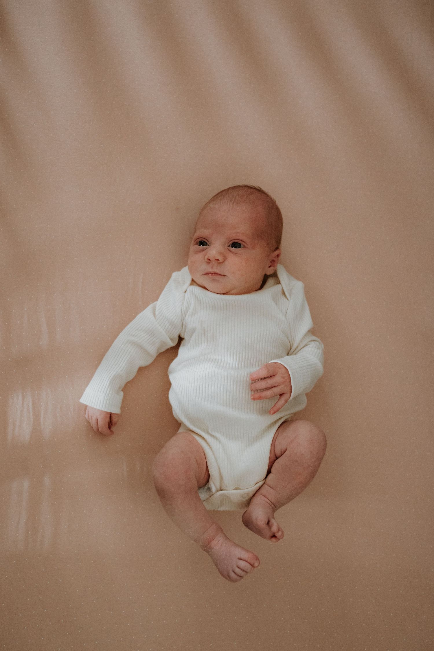 A baby wearing a white ribbed onesie lies on beige fabric in a series of gentle poses.