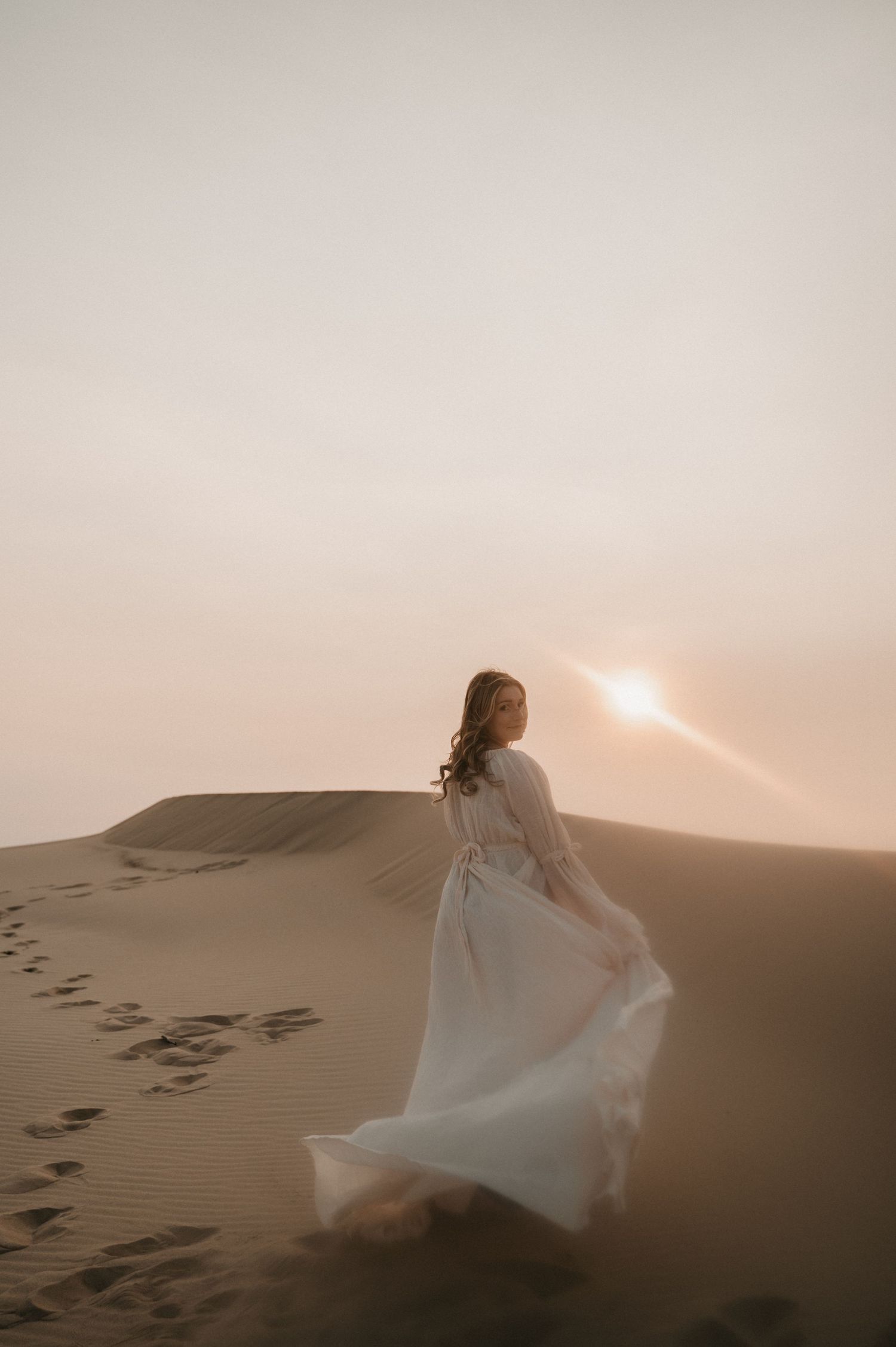 An ethereal white gown flows in the breeze atop desert sand dunes at sunset.