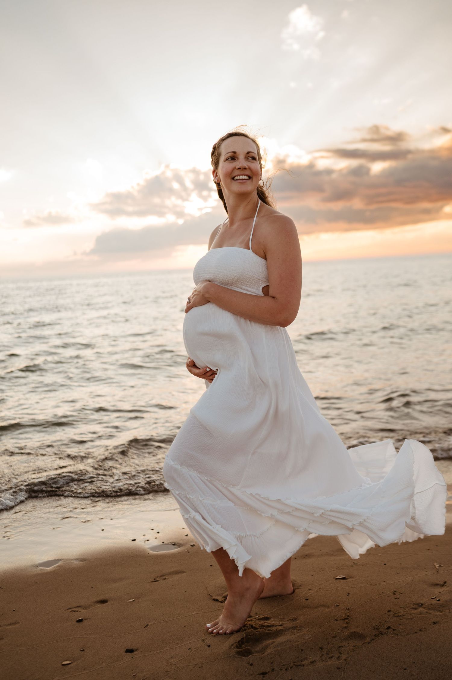 A pregnant woman in a flowing white dress twirls joyfully on the beach at sunset by the ocean.