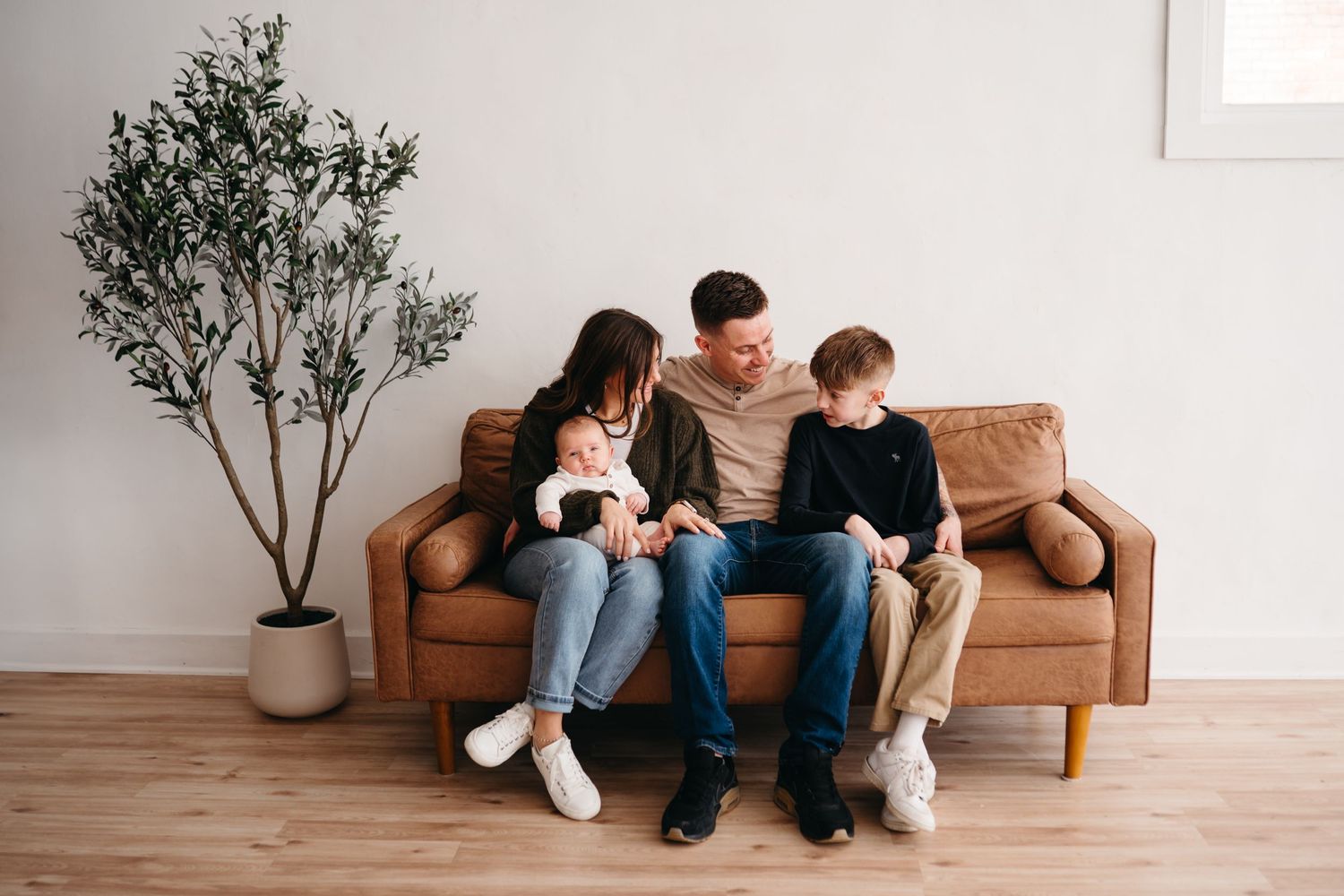 Family relaxing together on a brown leather couch in a minimalist living room with a potted olive tree.