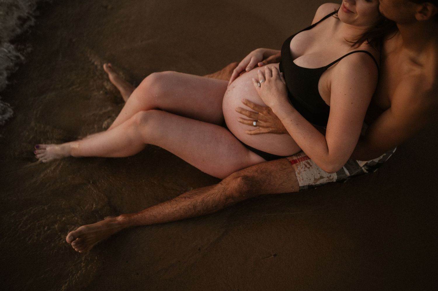 Intimate beach maternity portrait showing couple sitting in wet sand at golden hour.