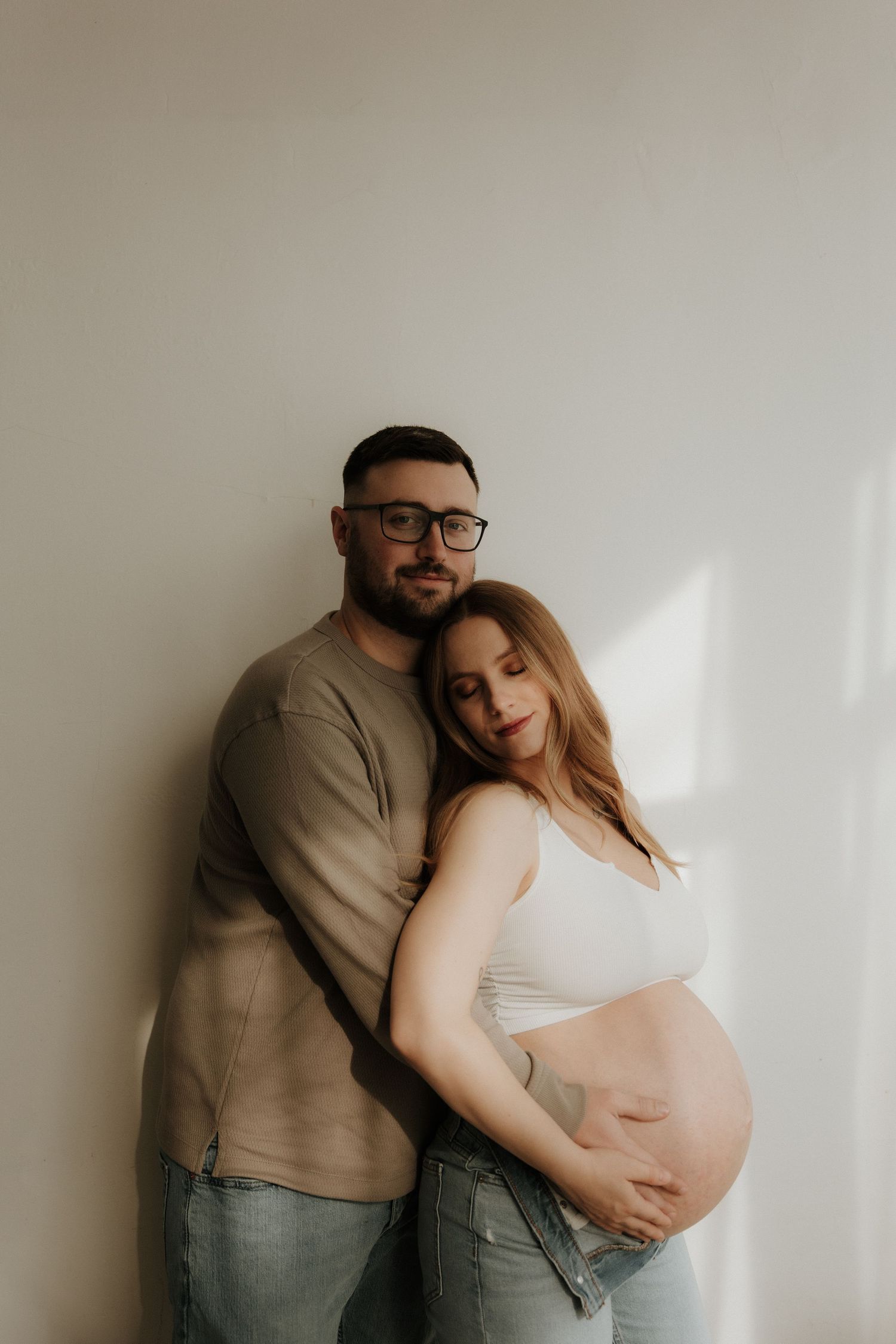 Intimate maternity photoshoot featuring a couple in casual attire against a white wall with natural window light.