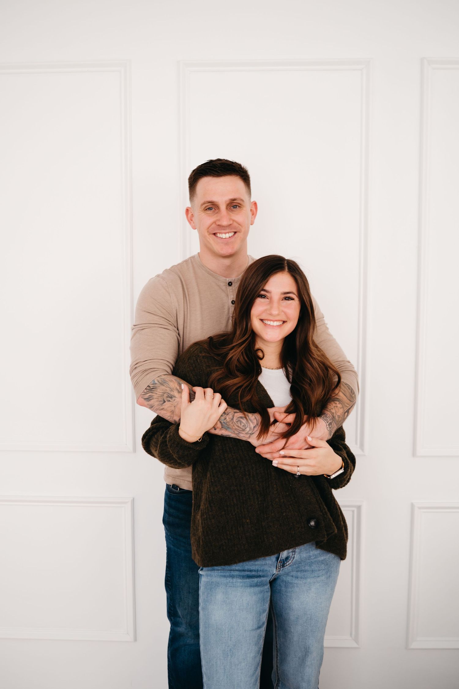 A happy couple embraces while standing against a white wall, wearing casual clothing and sharing genuine smiles.