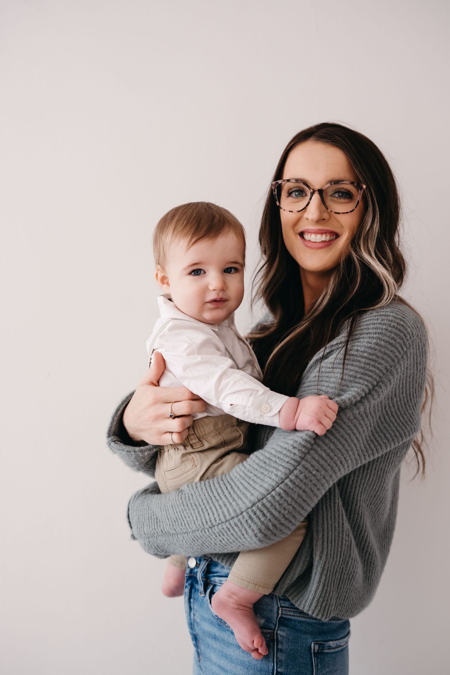 Mother in gray sweater holding baby in white outfit against neutral background.