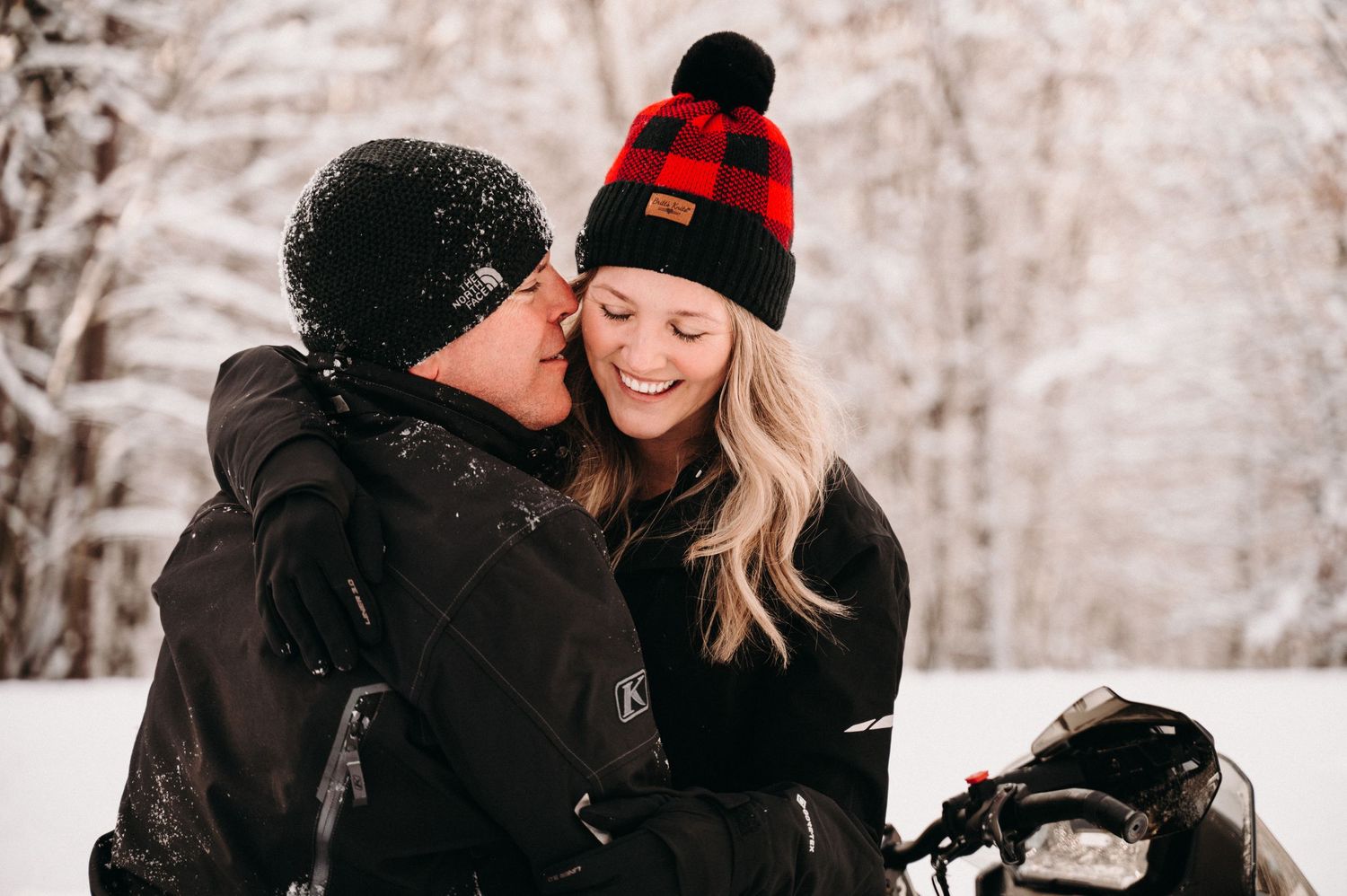 Couple in winter attire sharing intimate moment by snowmobile in snowy forest.