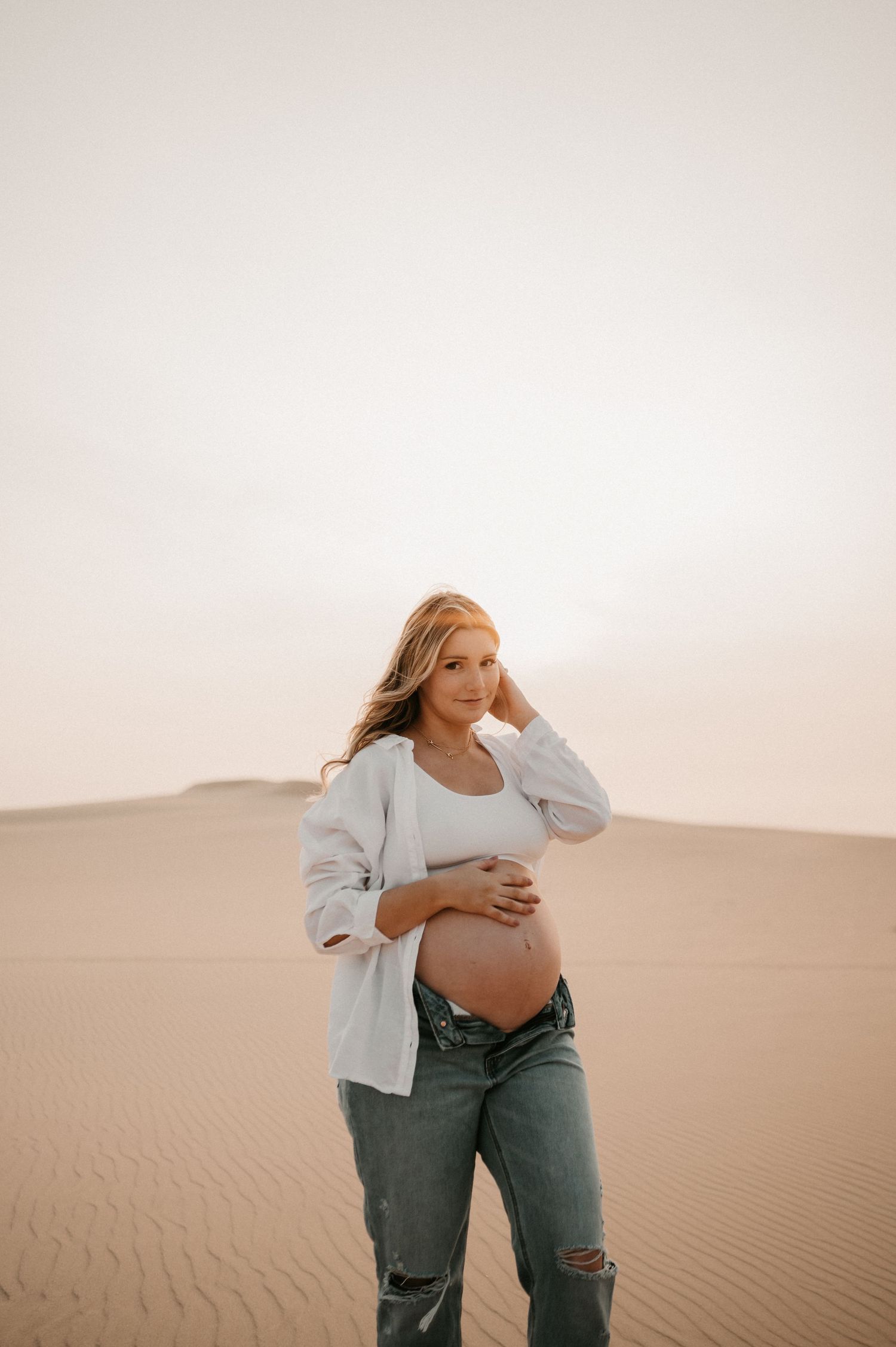 Maternity portrait of woman in casual attire standing among desert sand dunes.