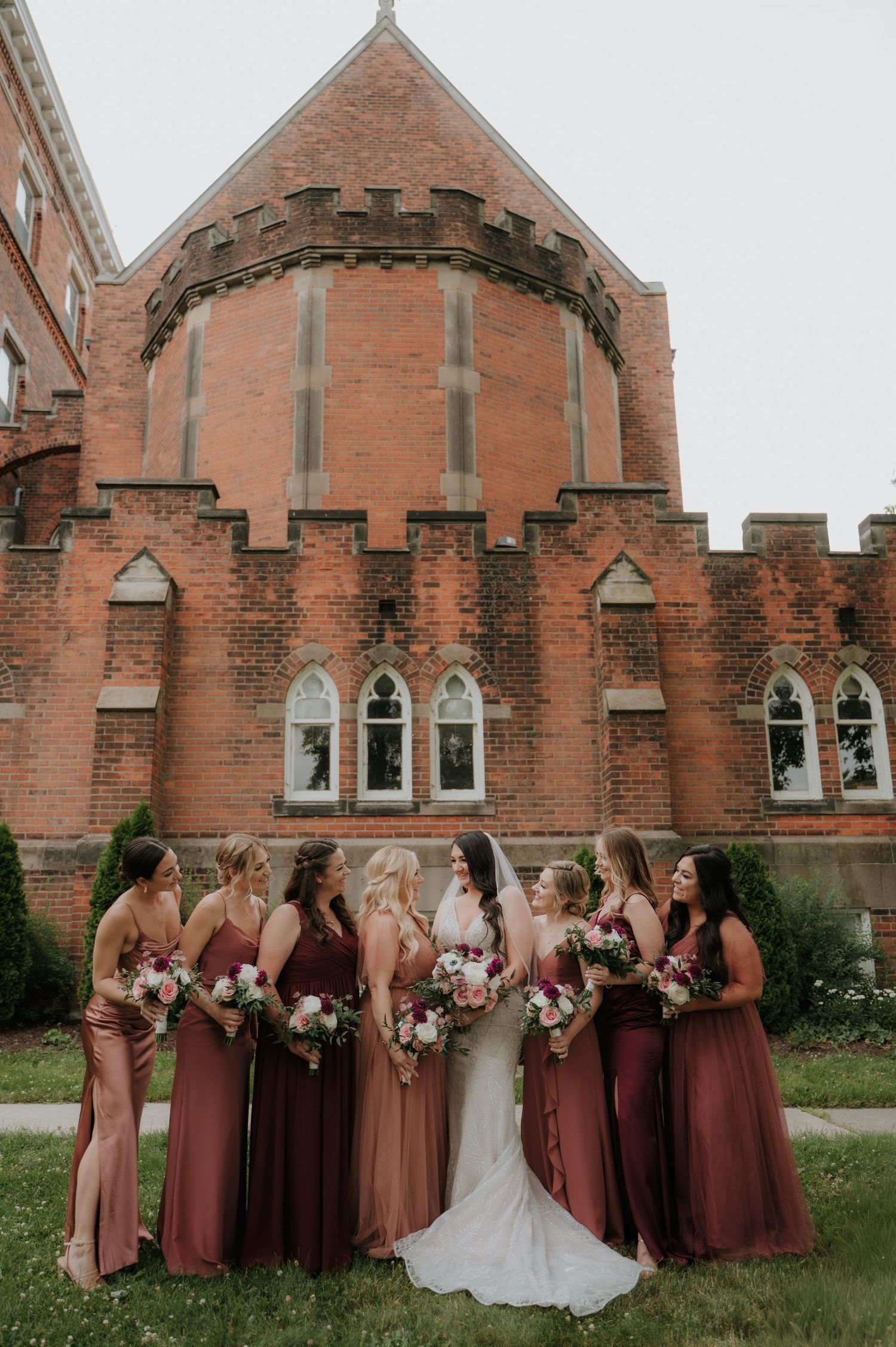 Wedding party poses in front of Gothic brick church wearing burgundy dresses.