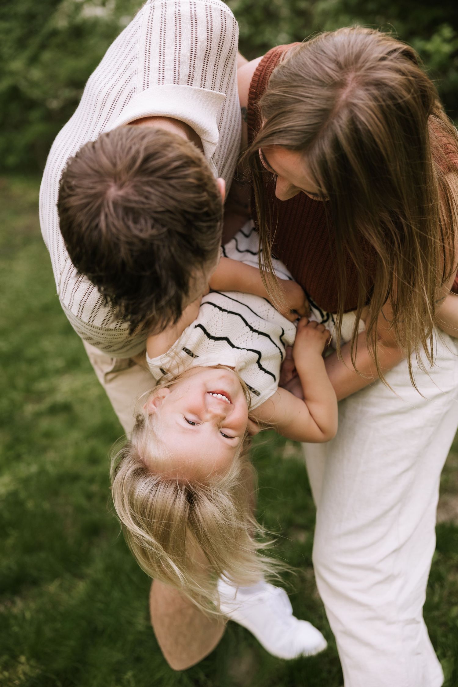 Two parents playfully hold their giggling toddler upside down together in a sunny green outdoor setting.