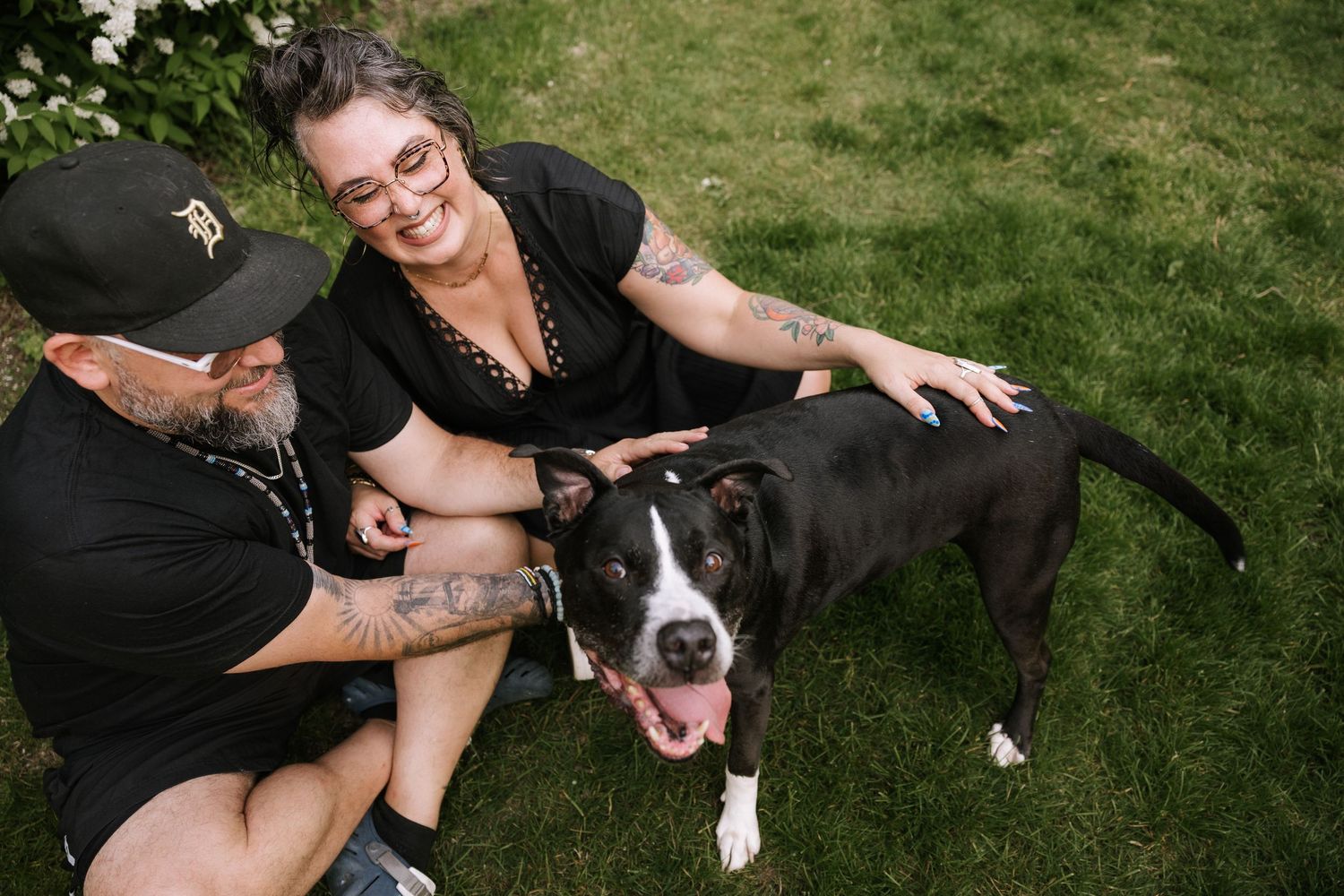 Smiling couple with tattoos sitting on grass with their black and white pit bull dog outdoors.