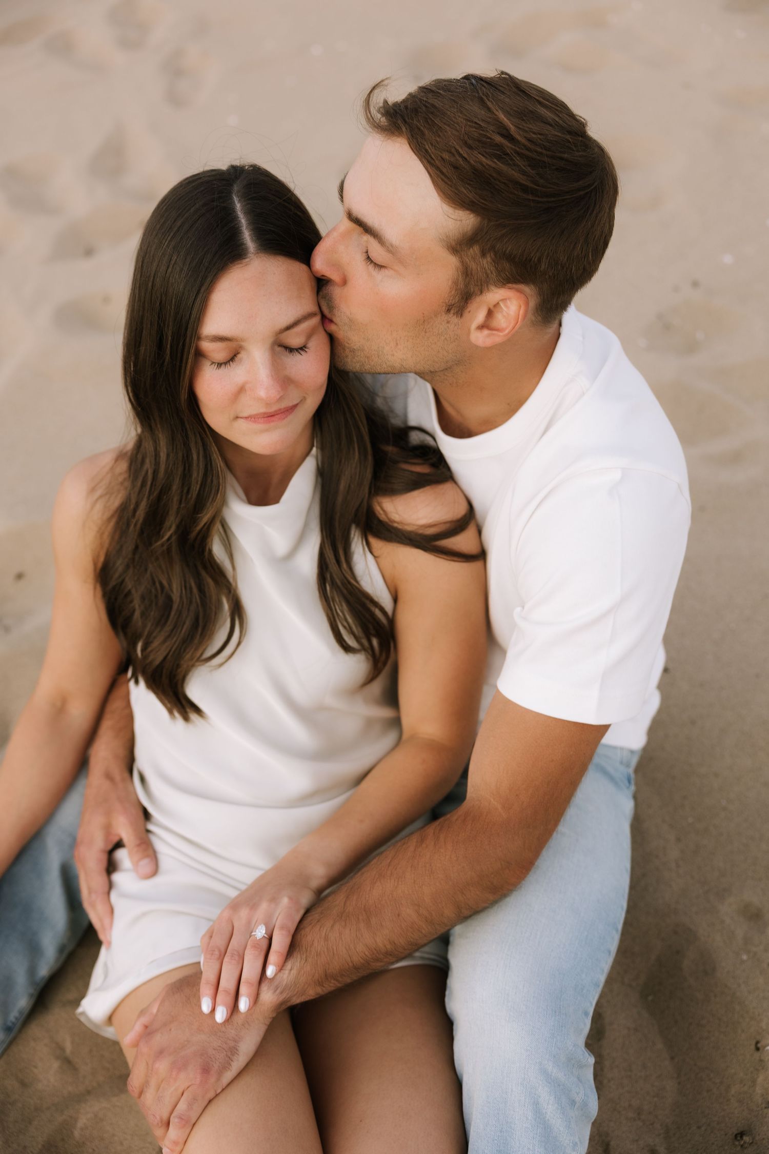 Man tenderly kissing womans cheek while sitting together on sandy beach, both wearing white outfits.