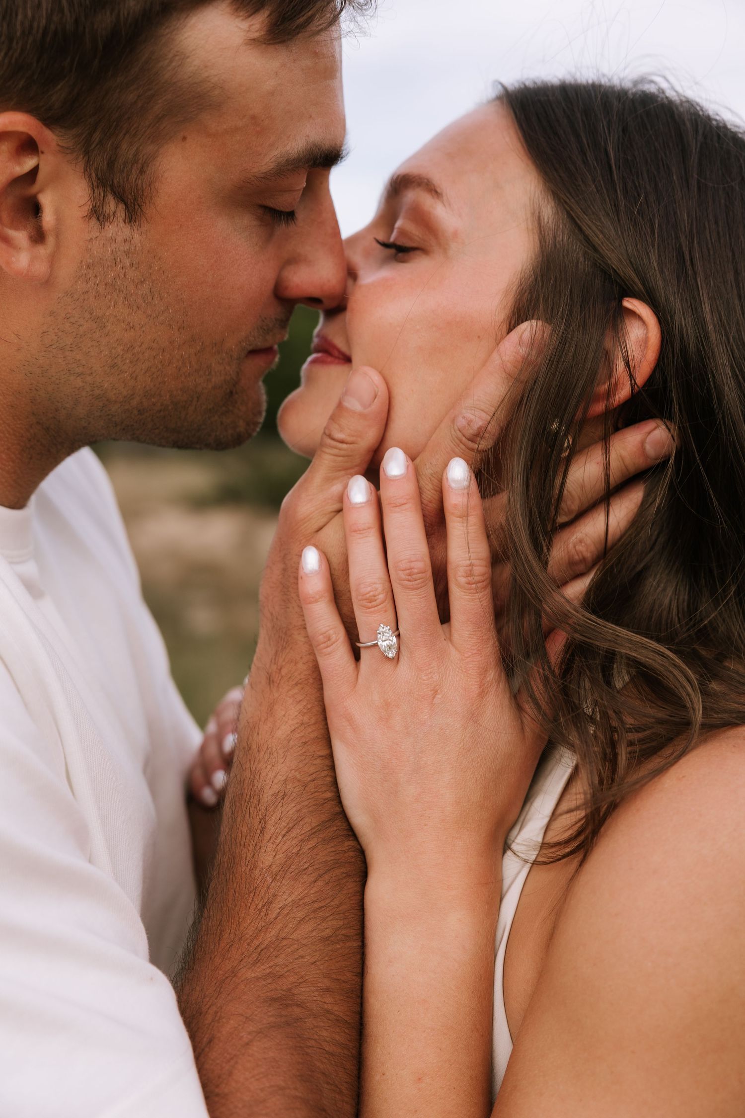 Couple sharing a romantic kiss, woman displaying elegant engagement ring while man tenderly holds her face outdoors.