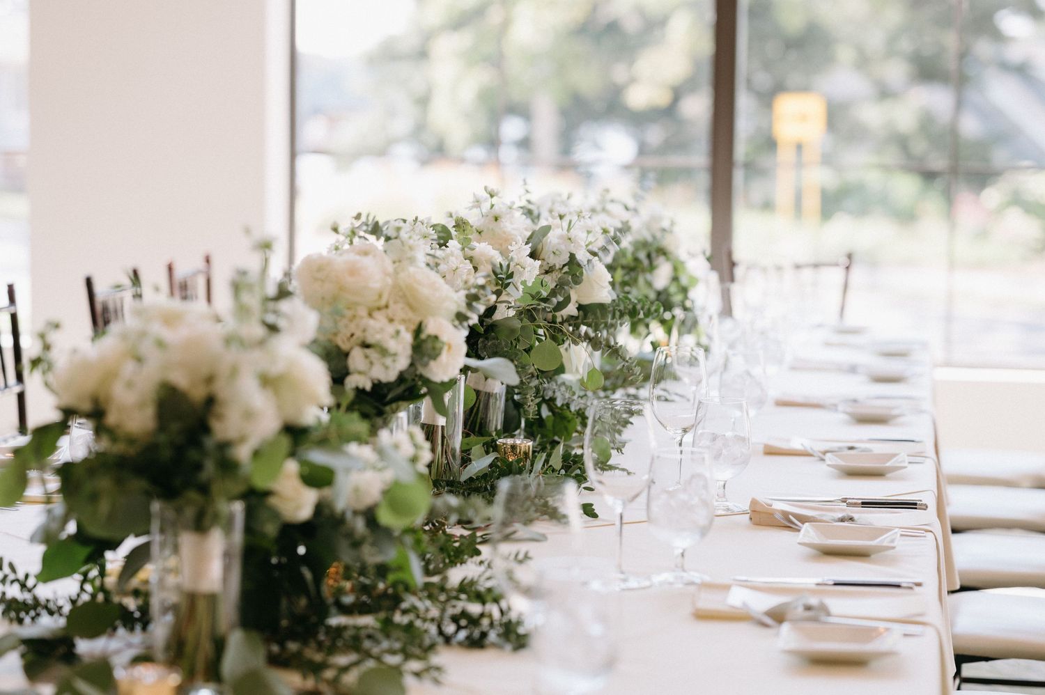 Elegant white floral centerpieces and table settings at a wedding reception with natural lighting.