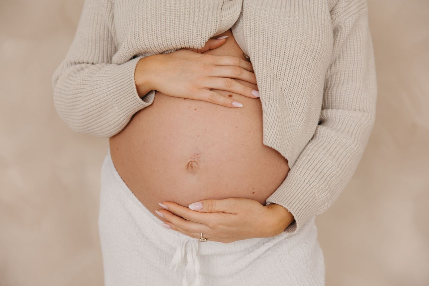 Close-up of pregnant woman in cream knit sweater gently holding bare baby bump with both hands.