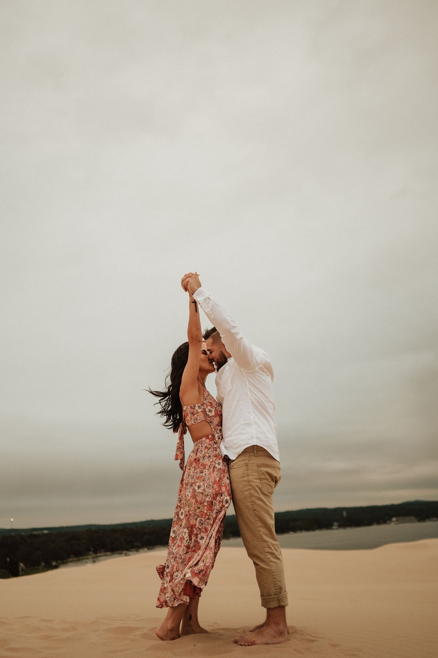 A couple dances together barefoot on sandy dunes against a moody sky during a romantic photoshoot.