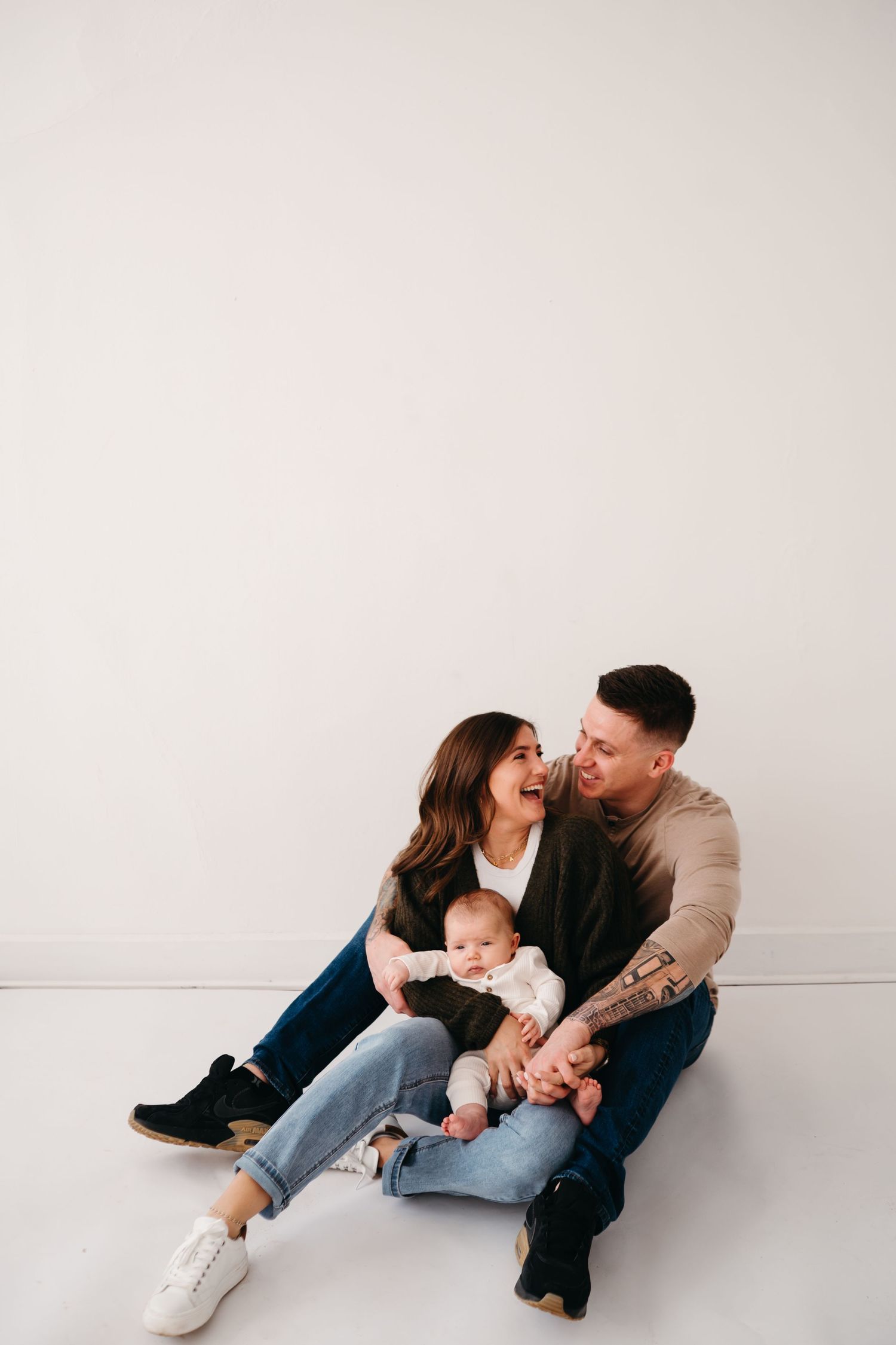 Family sits together on floor against white wall sharing intimate moment with their baby.