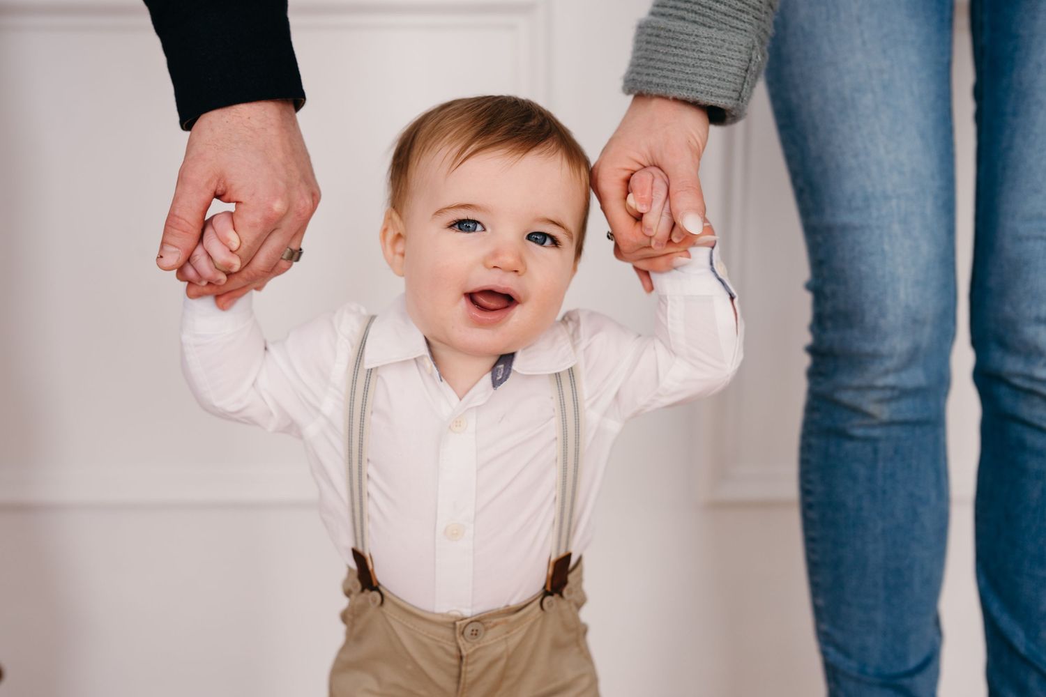Toddler in white shirt and tan suspenders smiles brightly while holding adult hands on both sides.