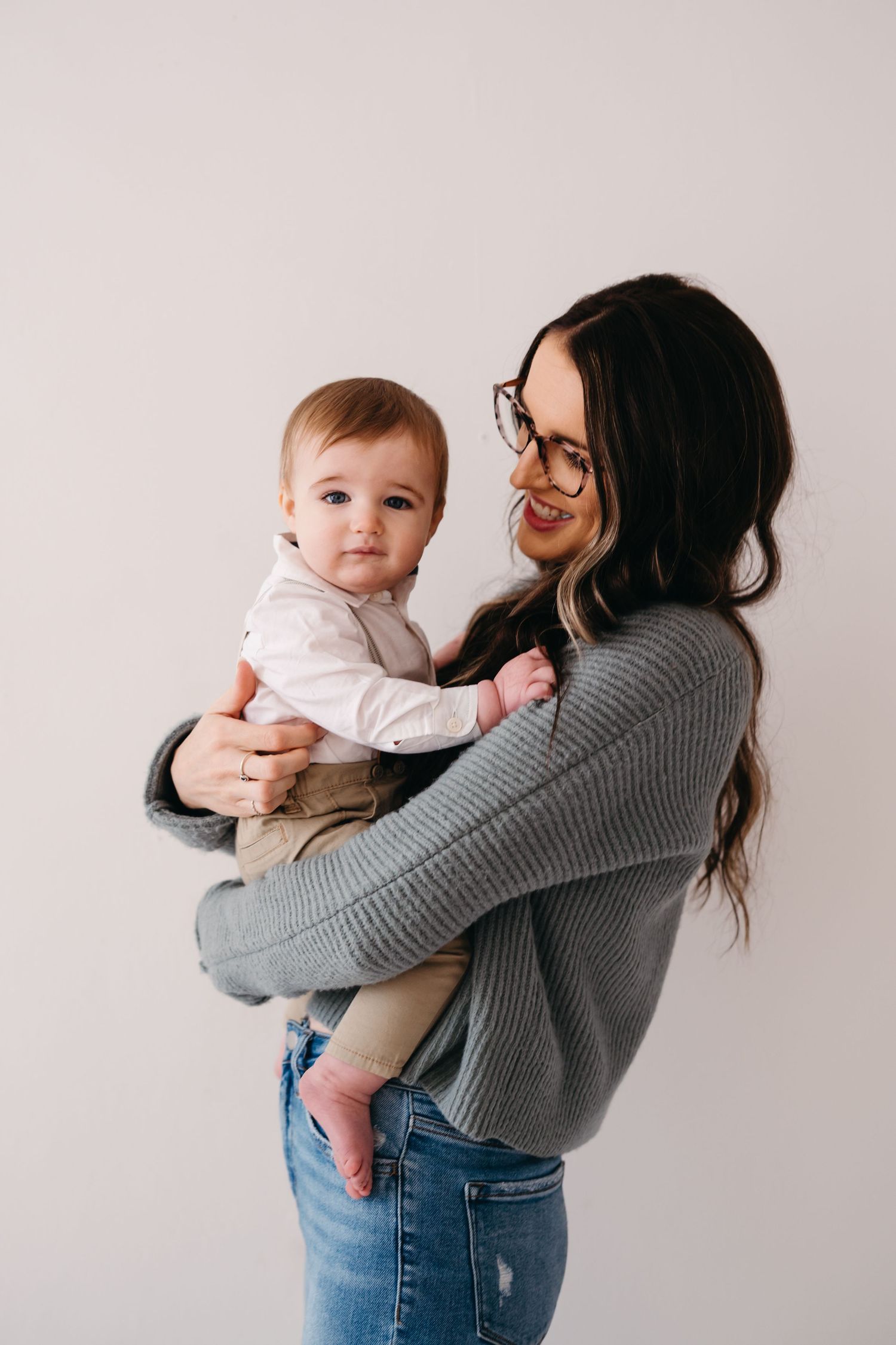 Woman in gray sweater and glasses holds baby wearing white shirt against light background.