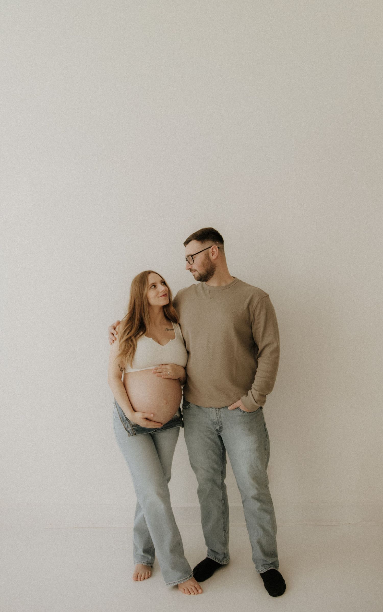 Minimalist maternity portrait of couple in casual attire standing against white backdrop.