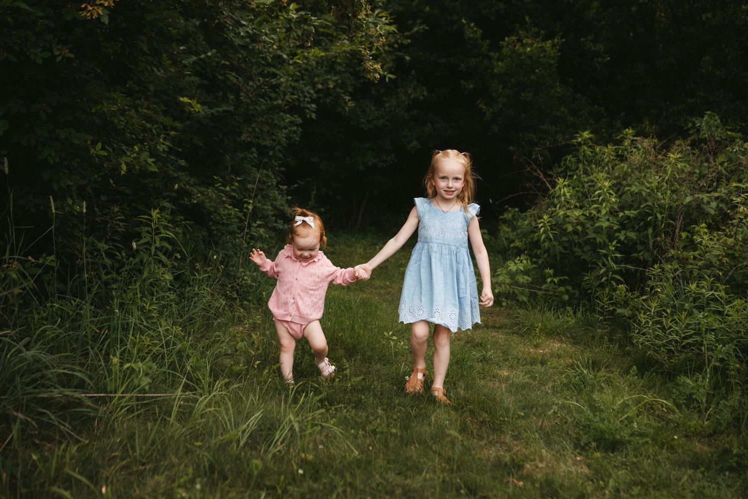 Two young children hold hands while running through a grassy outdoor path.