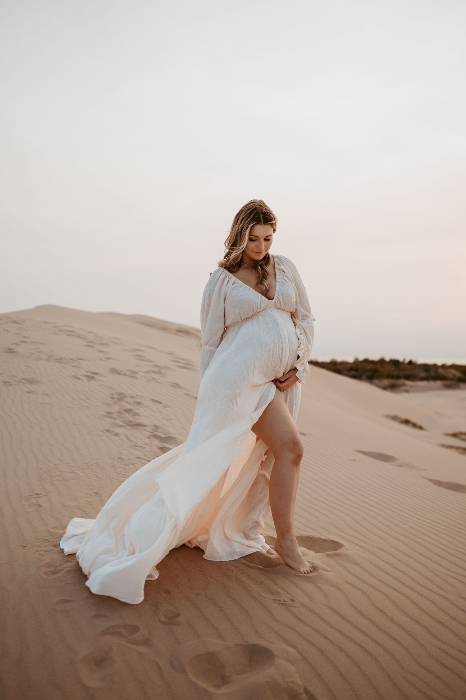 A woman in a flowing white dress poses on desert sand dunes at sunset.