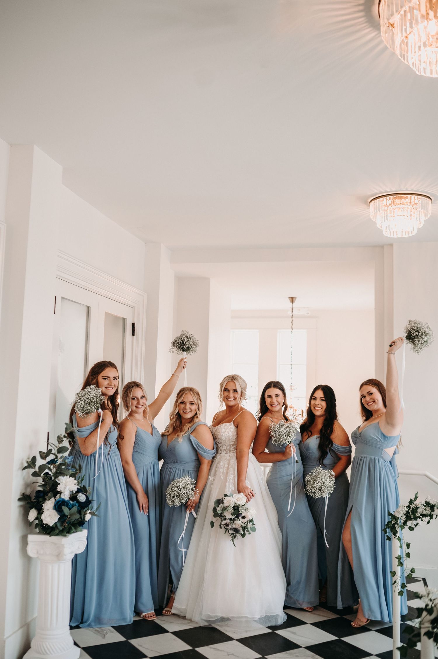 Wedding party celebrates joyfully in elegant white room with checkered floor and floral arrangements.