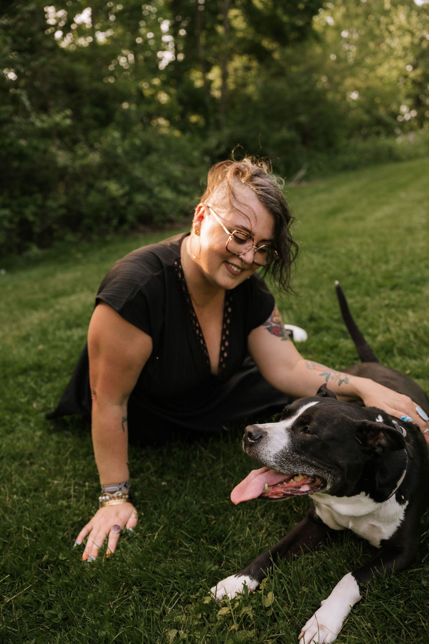 Woman with glasses and tattoos smiles while sitting on grass with a black and white dog.