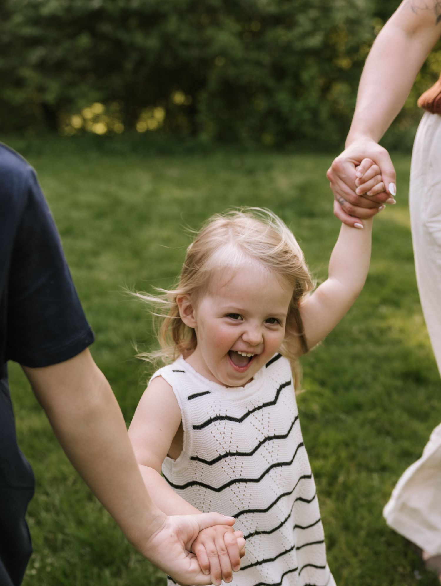 Joyful blonde toddler girl laughing while holding parents hands in green grassy park.