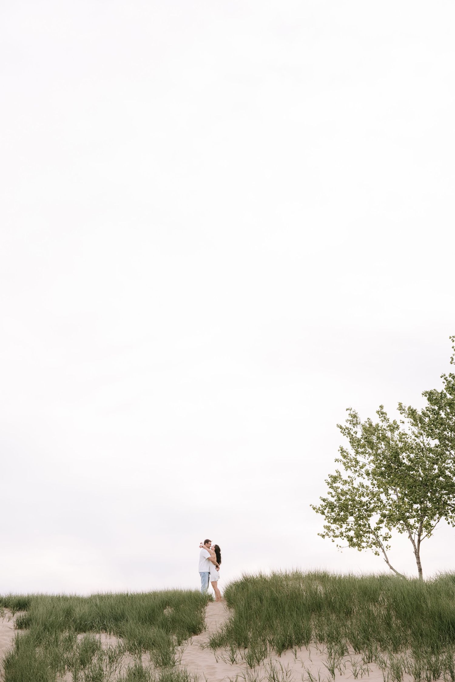 Couple embracing on a grassy sand dune beside a tree under an overcast sky.