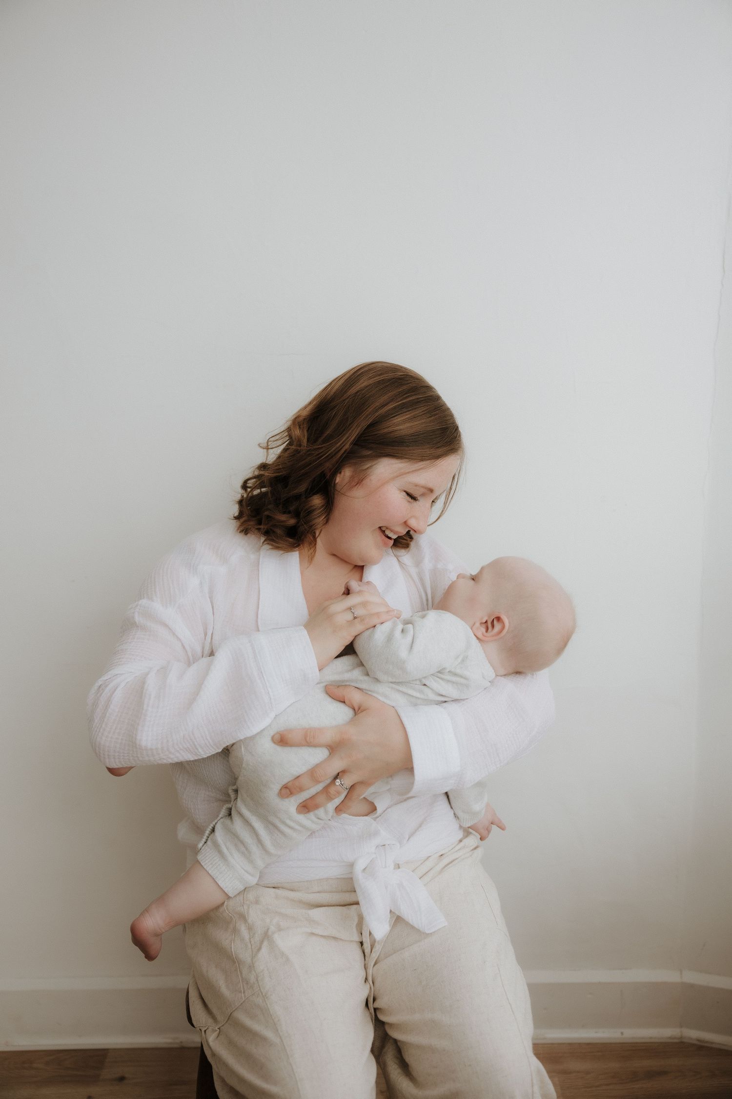 A smiling mother in white holds her newborn baby against a light background.
