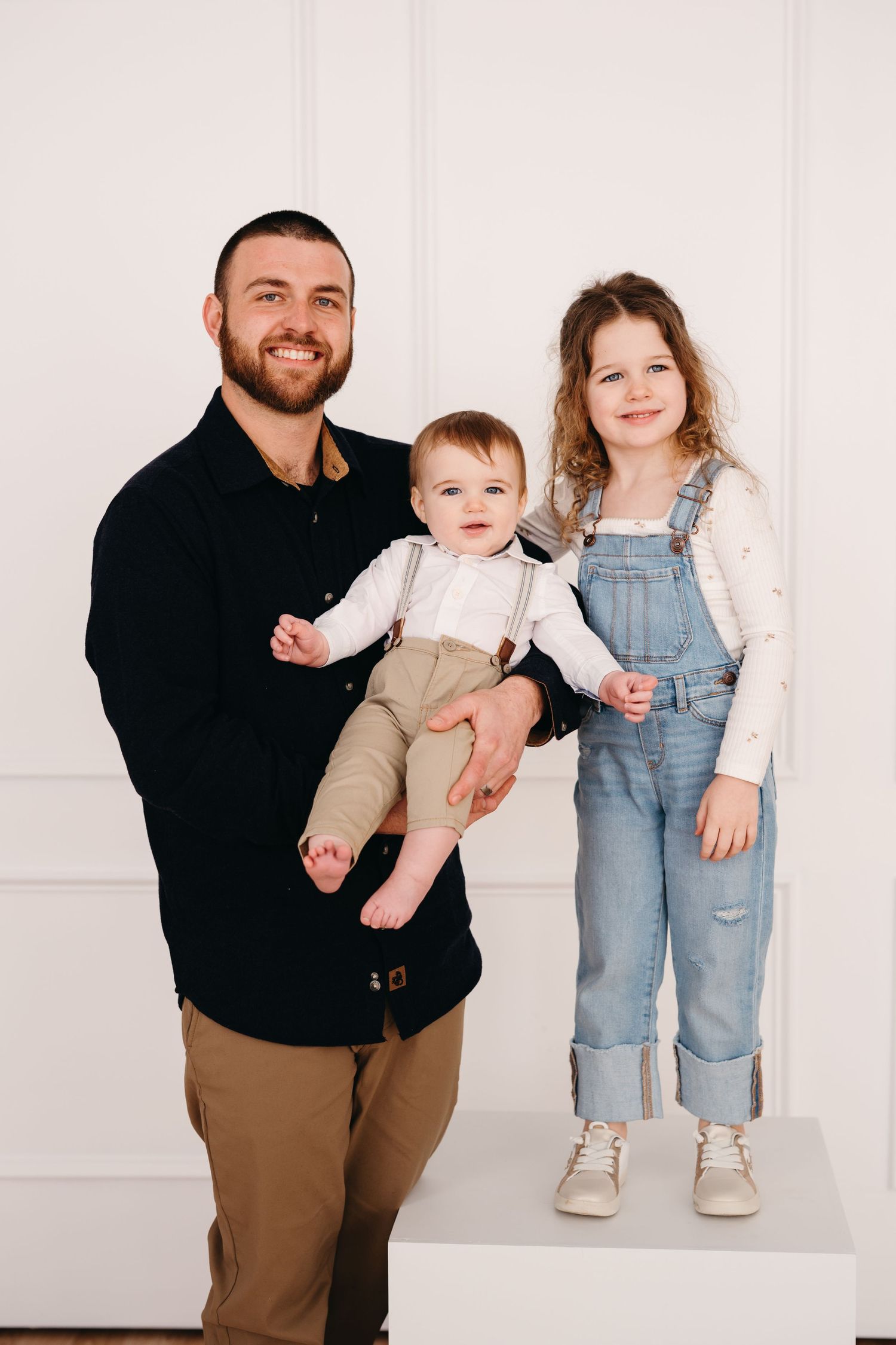 A family portrait against a white wall showing a parent in black holding a baby while a child stands beside in overalls.