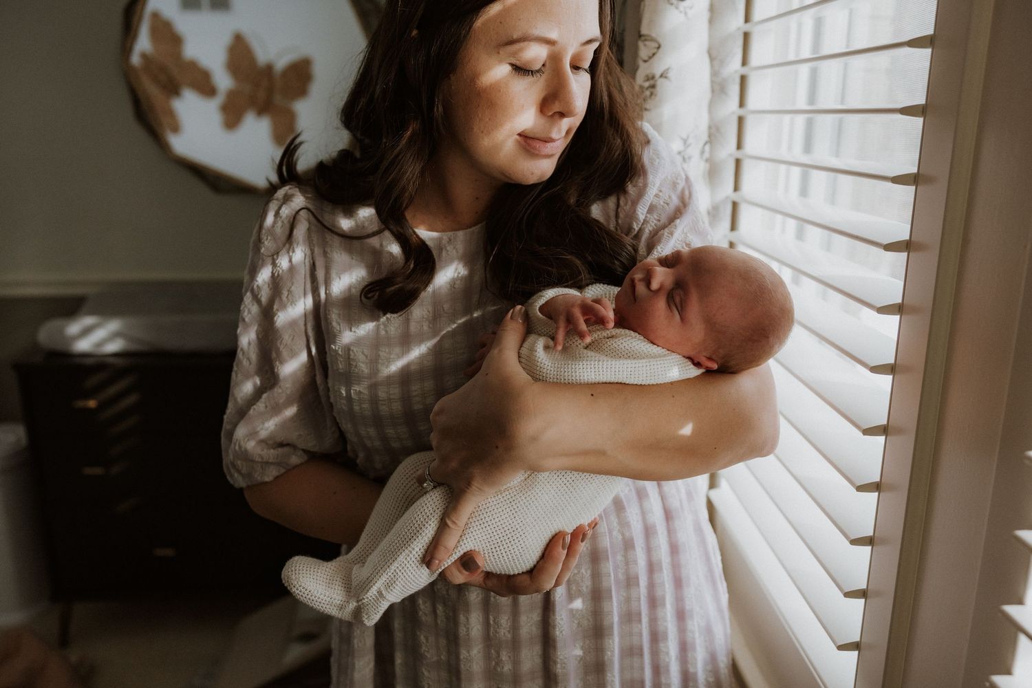 Mother holding newborn baby near window with soft natural lighting streaming through blinds.