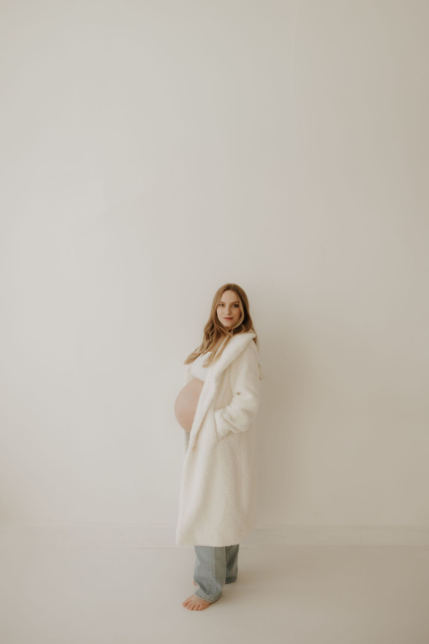 Maternity photos featuring woman in cream cardigan against white wall.