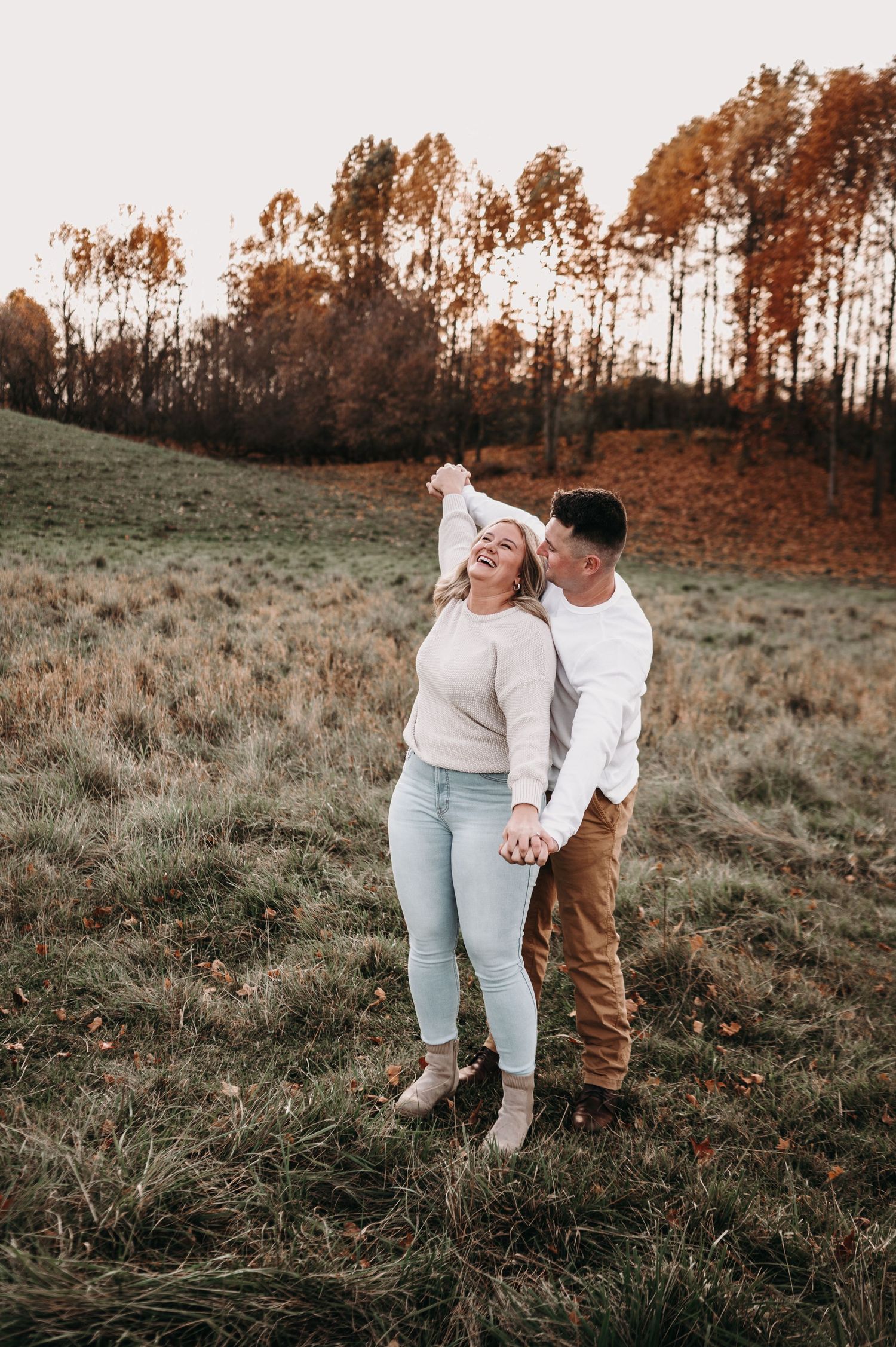 Couple shares a playful moment in a field during autumn sunset with tall trees in background.
