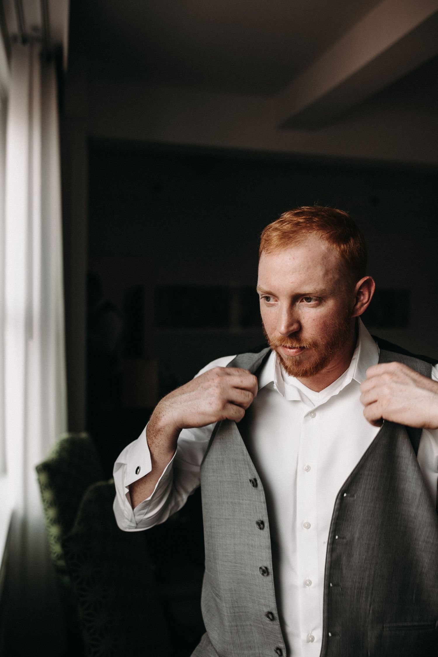 A person adjusts their grey suit jacket while standing near a window in moody lighting.
