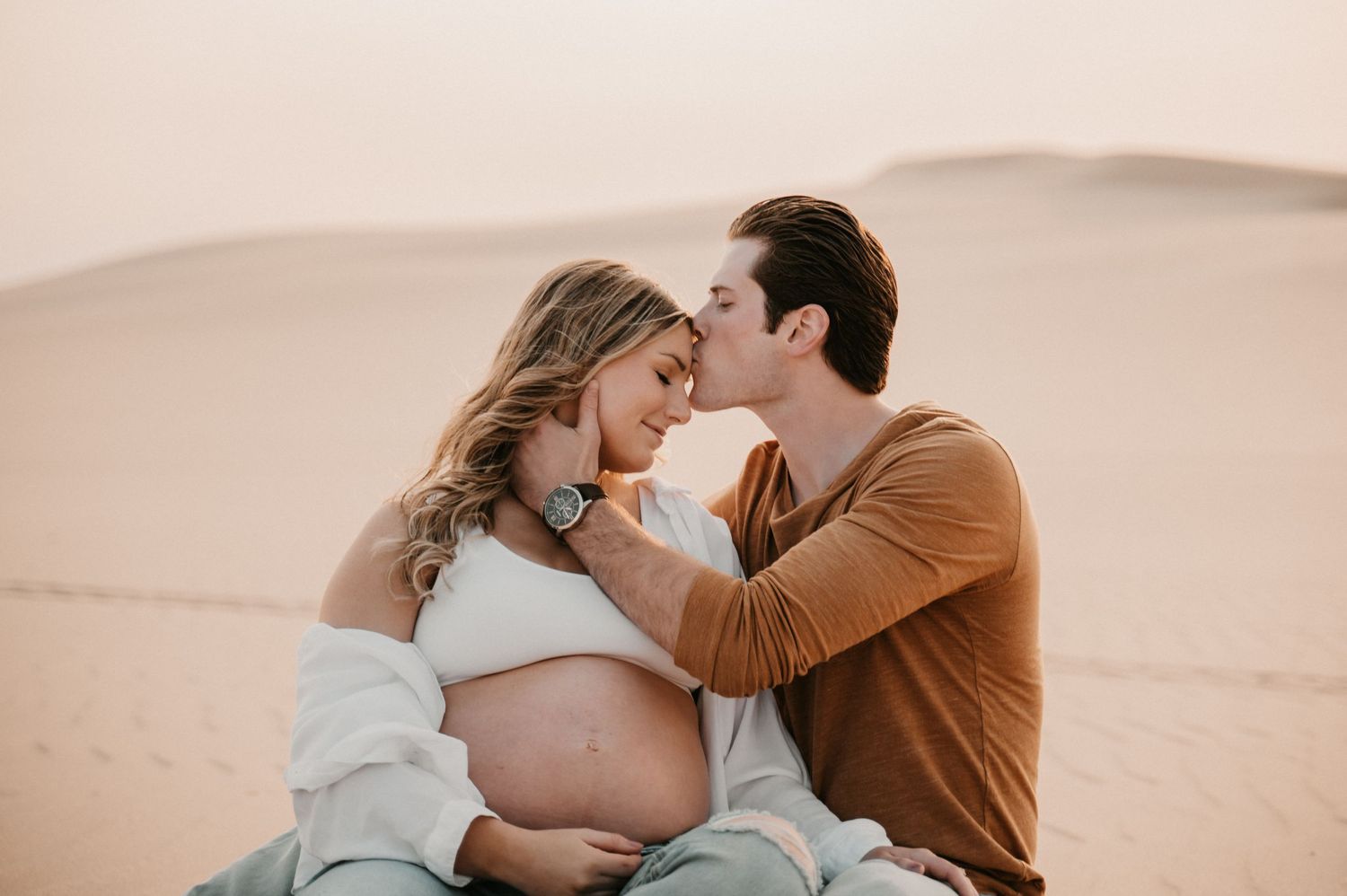 Intimate maternity photoshoot in desert setting with couple sitting on sand dunes at sunset.