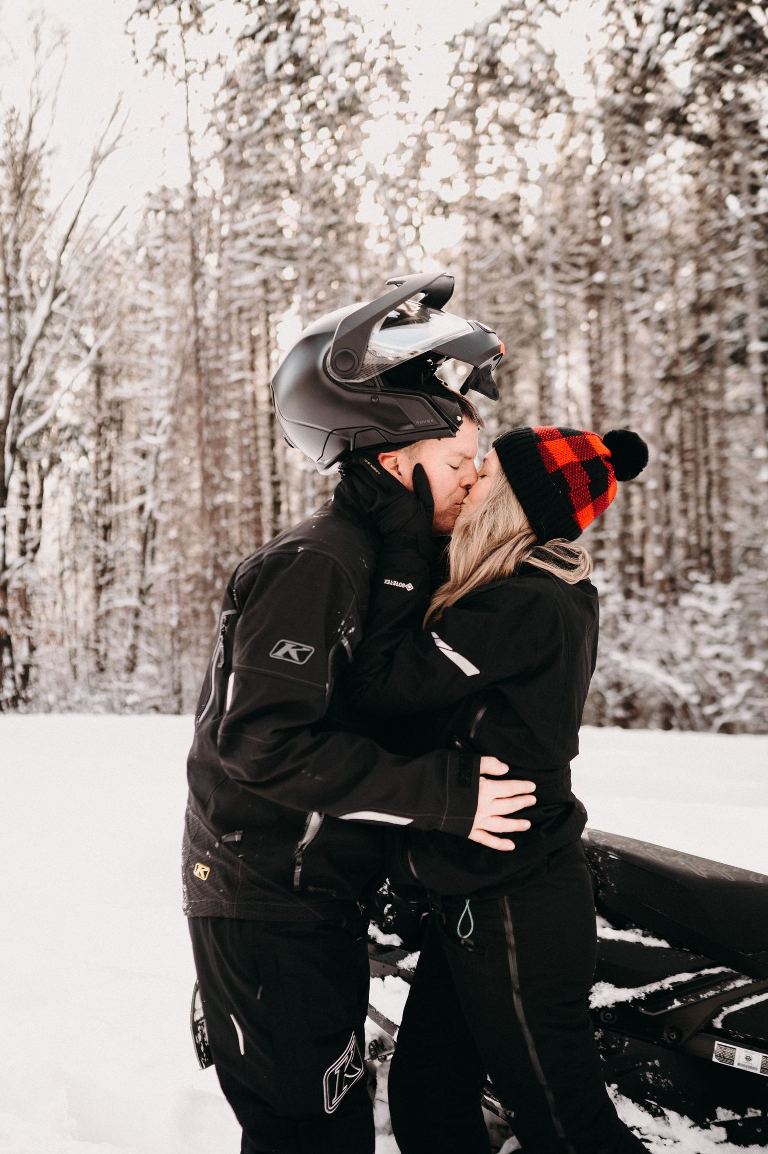 A couple in winter gear sharing a romantic moment next to a snowmobile in a snowy forest setting.
