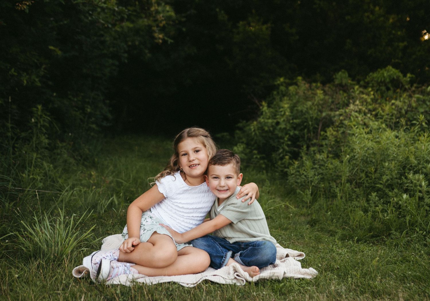 Two children sit together on a white blanket in a grassy outdoor setting.