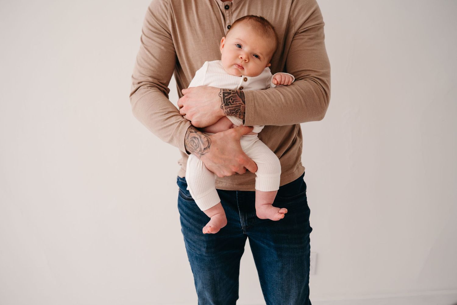 Baby in white onesie being held against beige sweater with denim jeans.