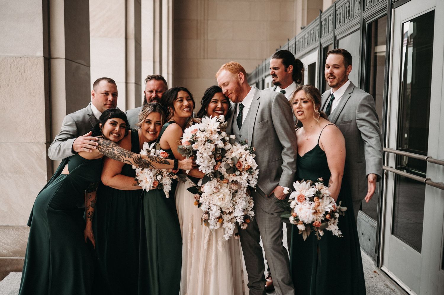 Bridal party laughs together while posing in formal wear against stone building backdrop.