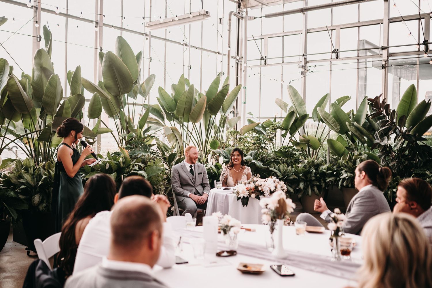 Wedding reception in a bright greenhouse with guests seated at decorated tables.