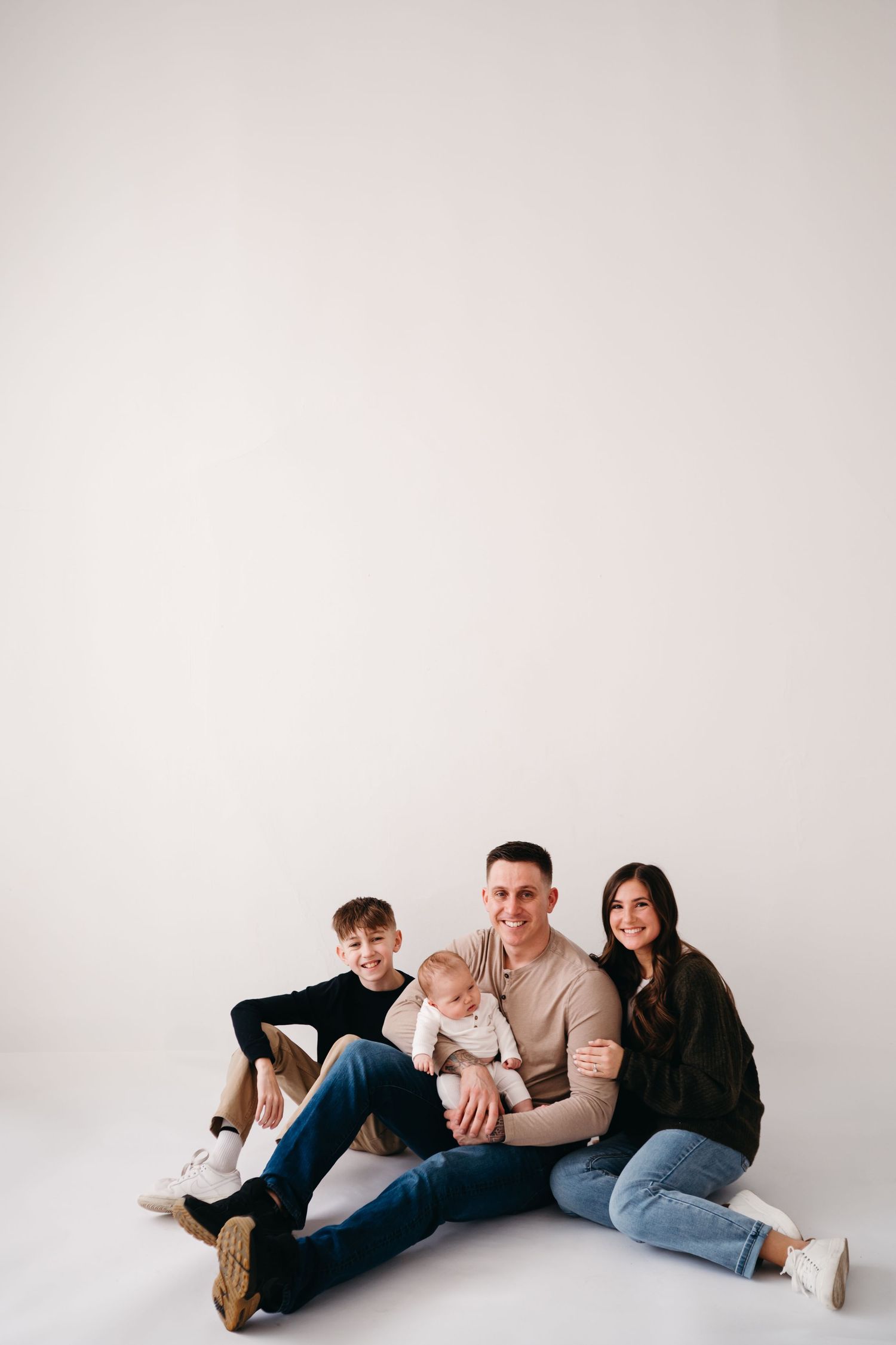 A family of four sits together on a white studio floor wearing casual clothing in a minimalist setting.