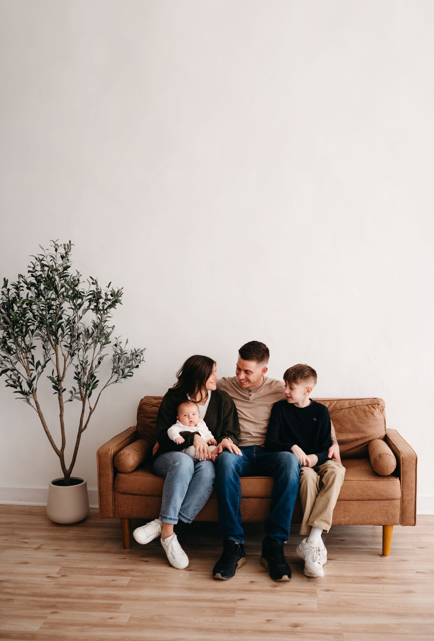 A family sits together on a brown leather couch next to a potted tree in a minimalist living room.