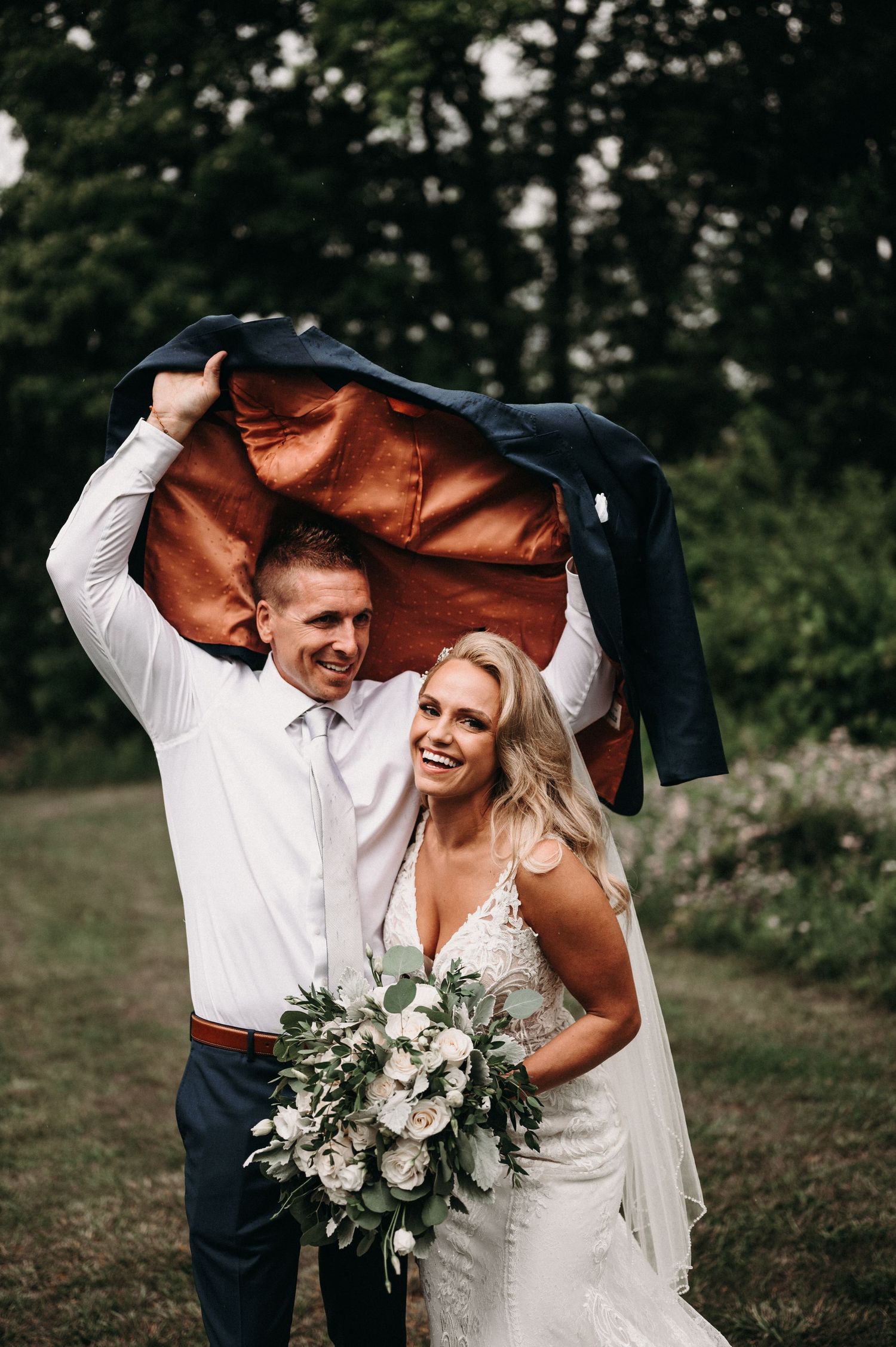 Newlyweds share a laugh while holding up a jacket for cover in a garden setting.