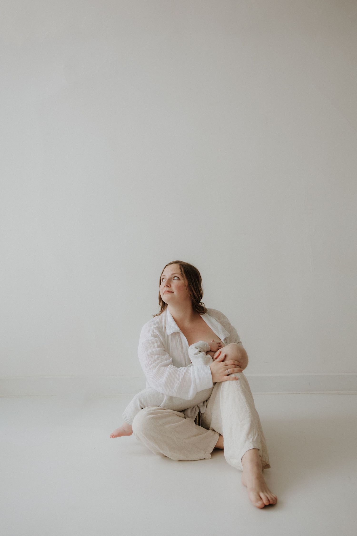 A woman in white linen sits on the floor, gazing upward thoughtfully in a bright, minimalist studio.