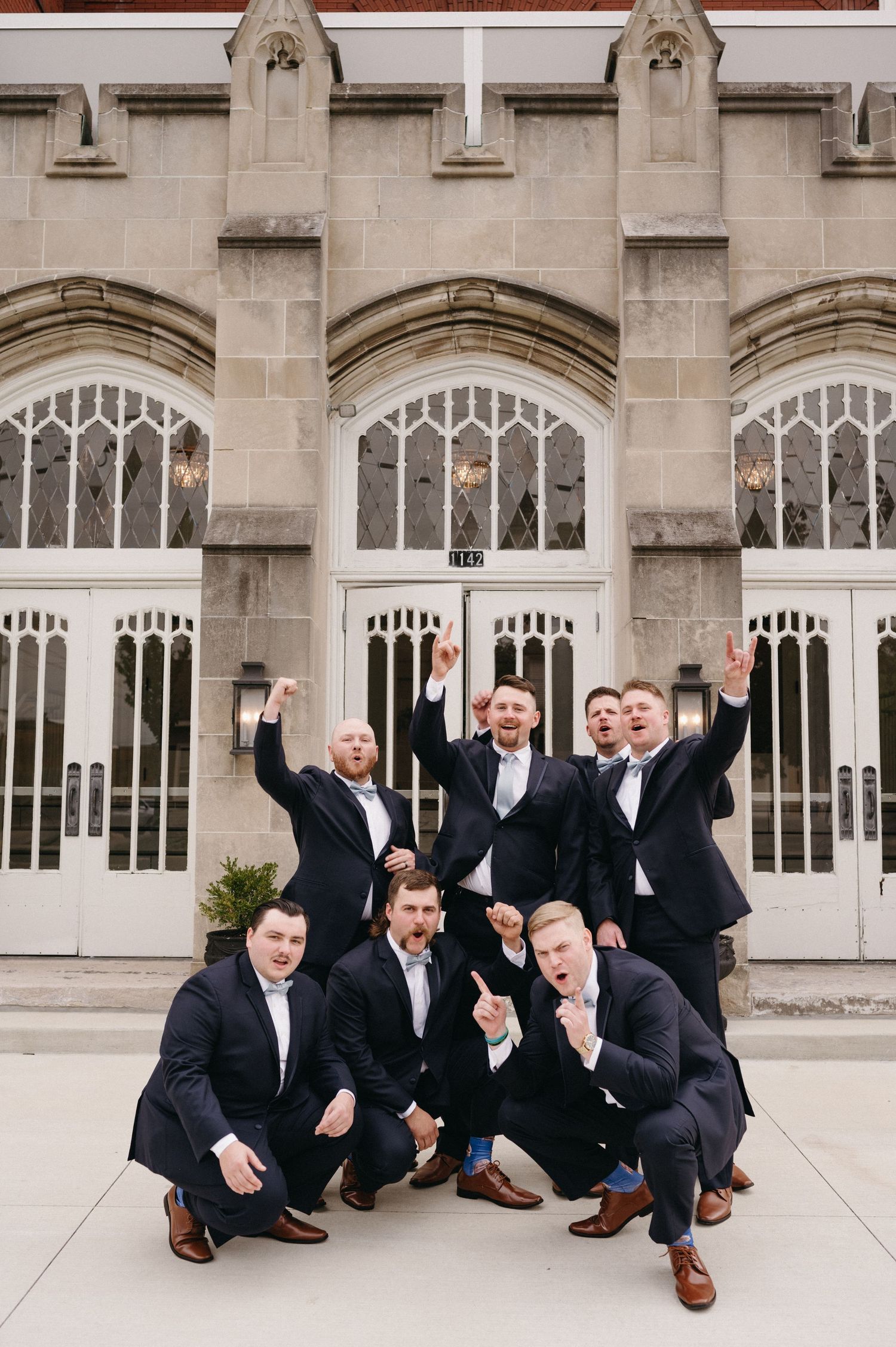 Wedding party of groomsmen striking fun poses in front of stone church facade.