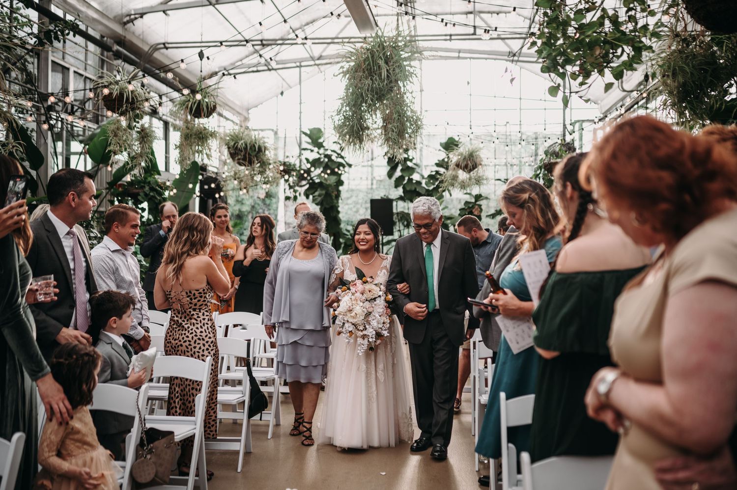 Wedding processional in a botanical greenhouse with guests lining the aisle.