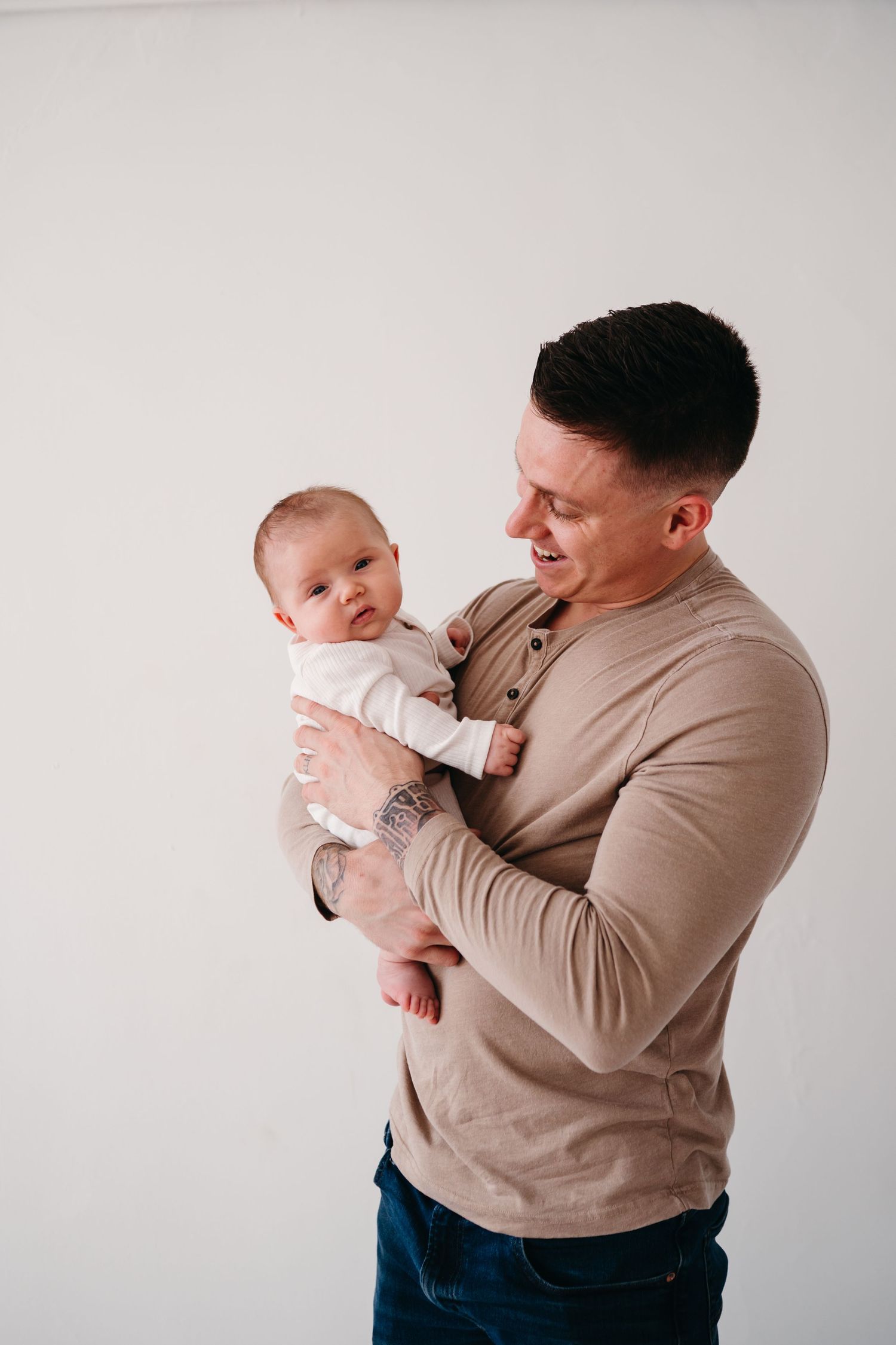 Parent wearing beige long sleeve shirt holds infant in white onesie against studio backdrop.