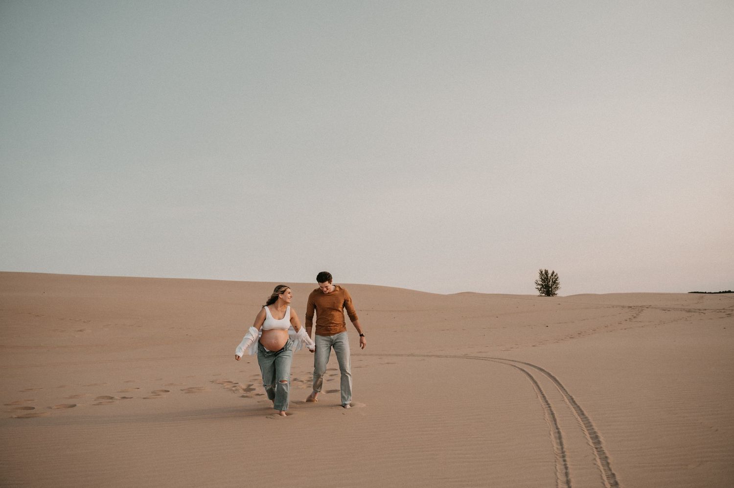 A couple walks hand in hand through desert sand dunes leaving footprints behind.