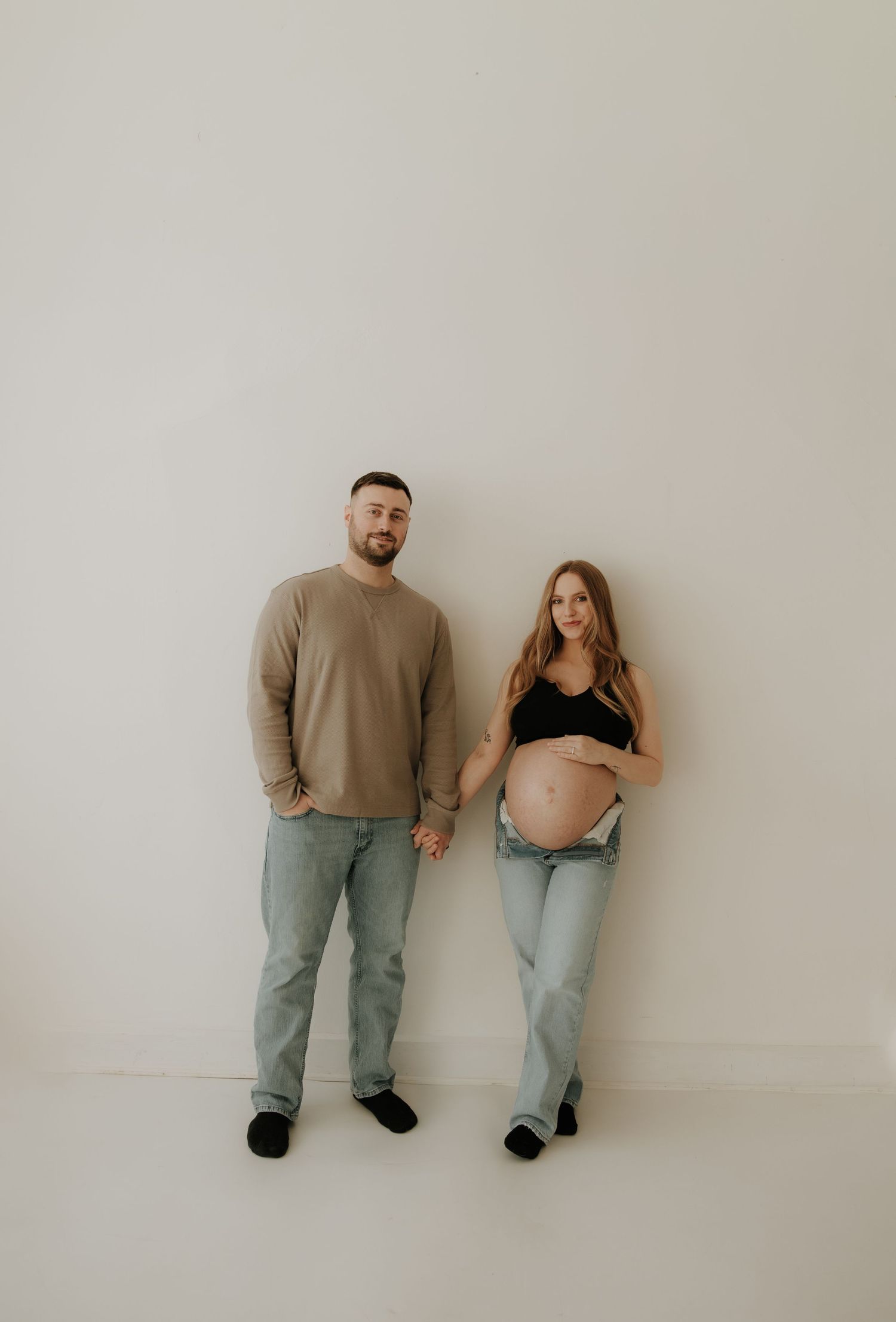 Full-length studio maternity photo of couple in casual wear against white wall.