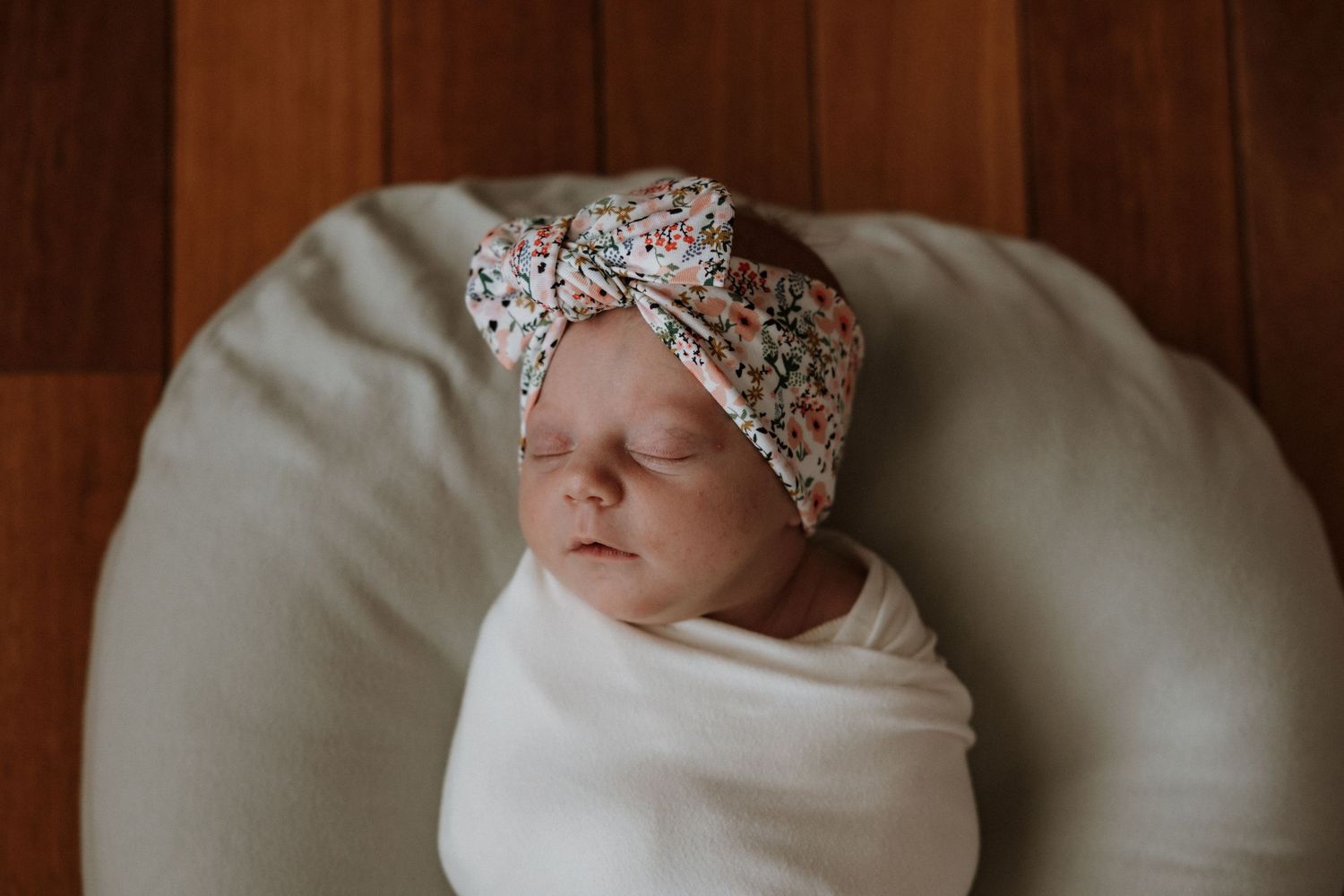 Sleeping newborn wrapped in white swaddle with floral headband on beige cushion.